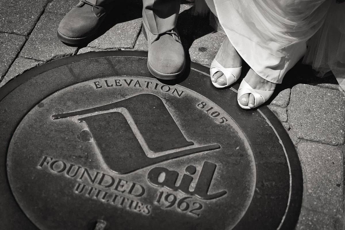 Black and white photo of a couple's feet on a "Vail Utilities" manhole cover. Bride wearing sandals, groom in shoes, suggesting a wedding setting.