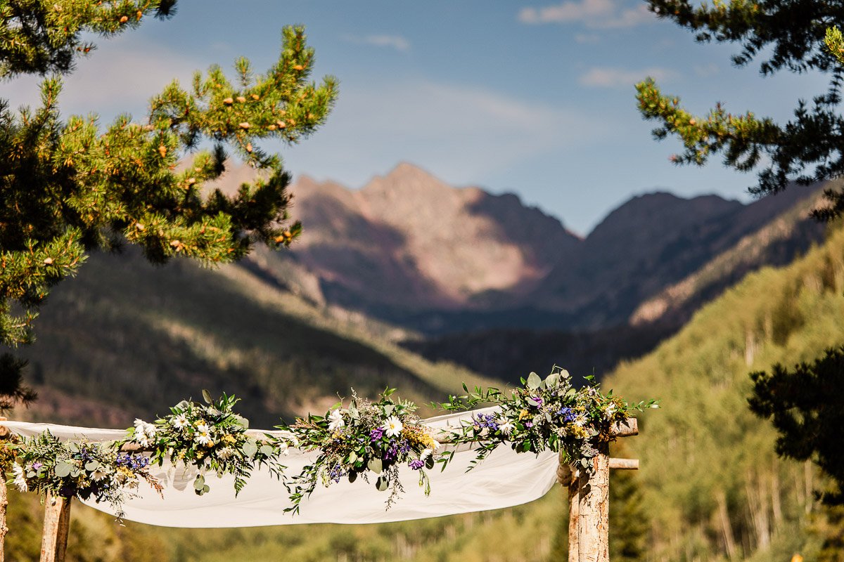 Rustic wedding arch adorned with white drapery and purple flowers, set against a backdrop of forested mountains and a clear blue sky. Tranquil ambiance.