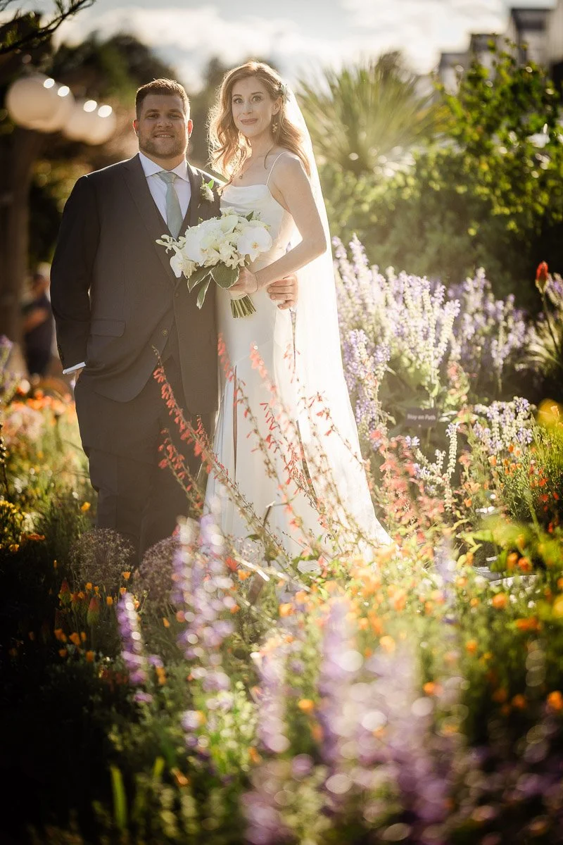 A bride and groom stand in a lush garden filled with colorful flowers. The bride holds a bouquet and wears a long, white dress, with soft sunlight enhancing the romantic scene.