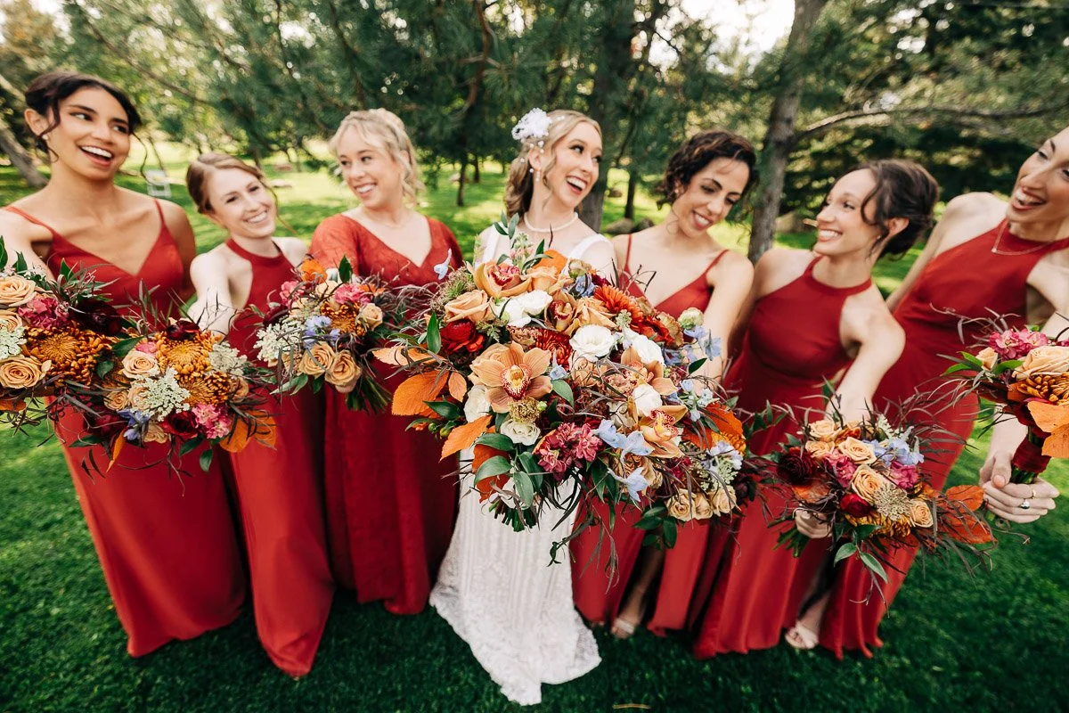 A joyful bride and six bridesmaids in red dresses hold vibrant autumn bouquets, smiling and laughing in a lush, green outdoor setting.