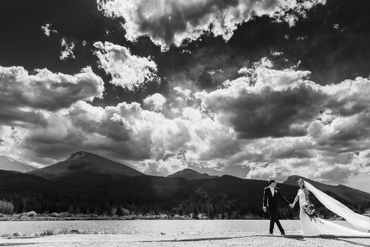 A bride and groom walk hand in hand against a dramatic backdrop of mountains and clouds, conveying romance and grandiosity. The bride’s veil flows elegantly.