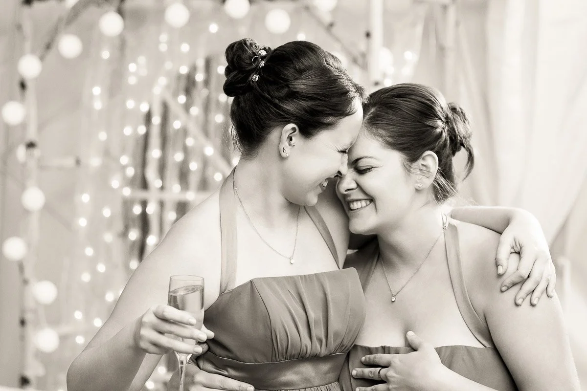 Two women in formal dresses share a joyful embrace, smiling with their foreheads touching. One holds a champagne glass. Lights twinkle softly in the background.