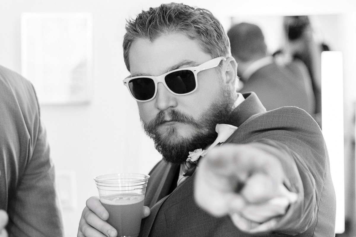 A man in sunglasses and a suit points playfully at the camera while holding a drink. The setting appears to be a lively event, suggesting a fun atmosphere.
