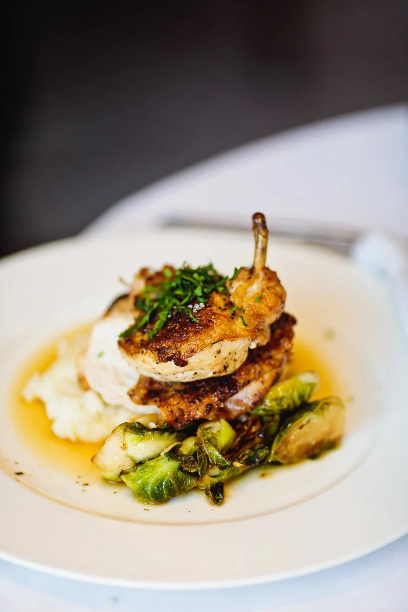 A gourmet dish with crispy roasted chicken atop mashed potatoes, garnished with herbs, served with sautéed Brussels sprouts on a white plate.