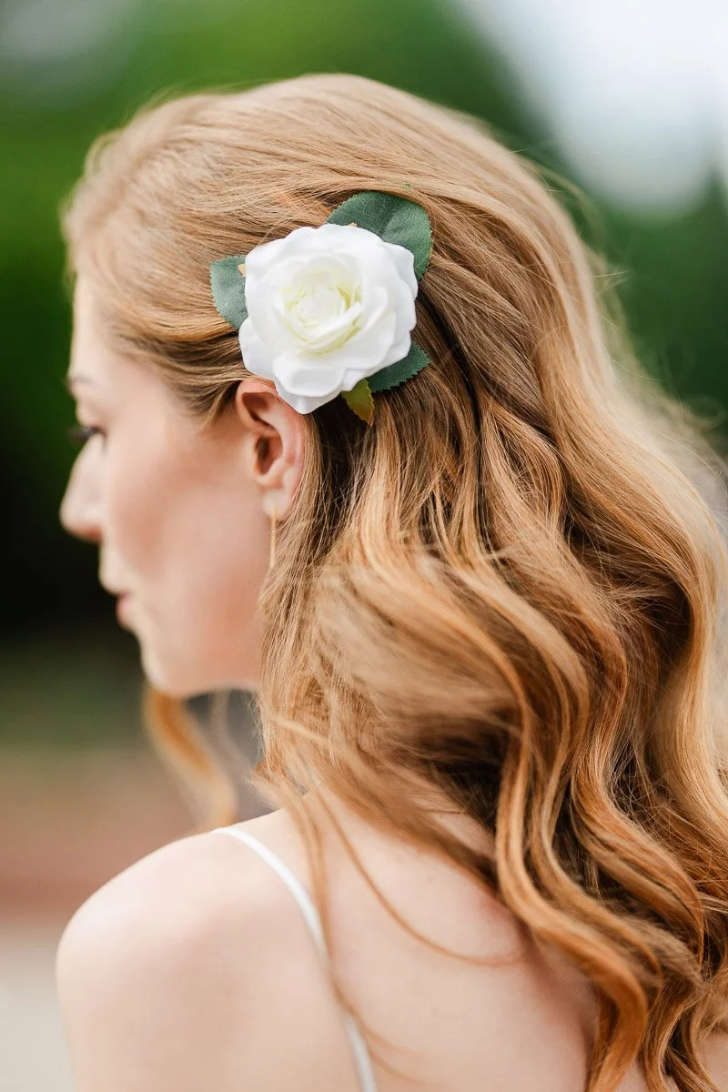 Profile of a woman with wavy blonde hair, adorned with a white rose clip. Soft focus and greenery create a serene, elegant atmosphere.