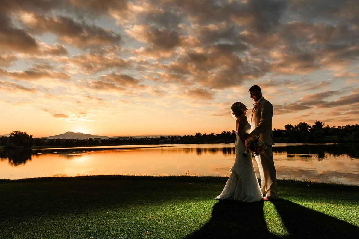 Silhouetted couple in wedding attire stands by a tranquil lake at sunset. The sky is golden with clouds, creating a romantic and serene atmosphere.