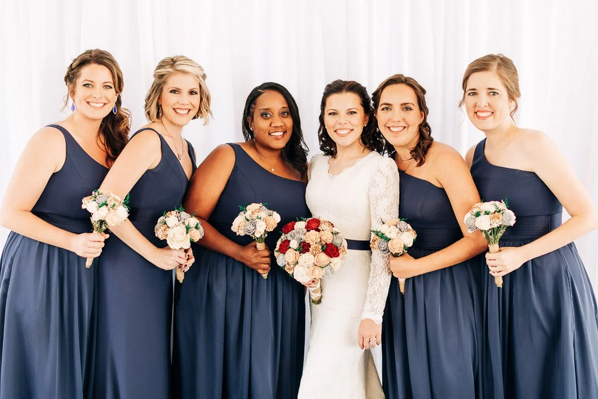 A bride in a white lace dress smiles with five bridesmaids in navy dresses holding bouquets. The scene is joyful and elegant against a white backdrop.