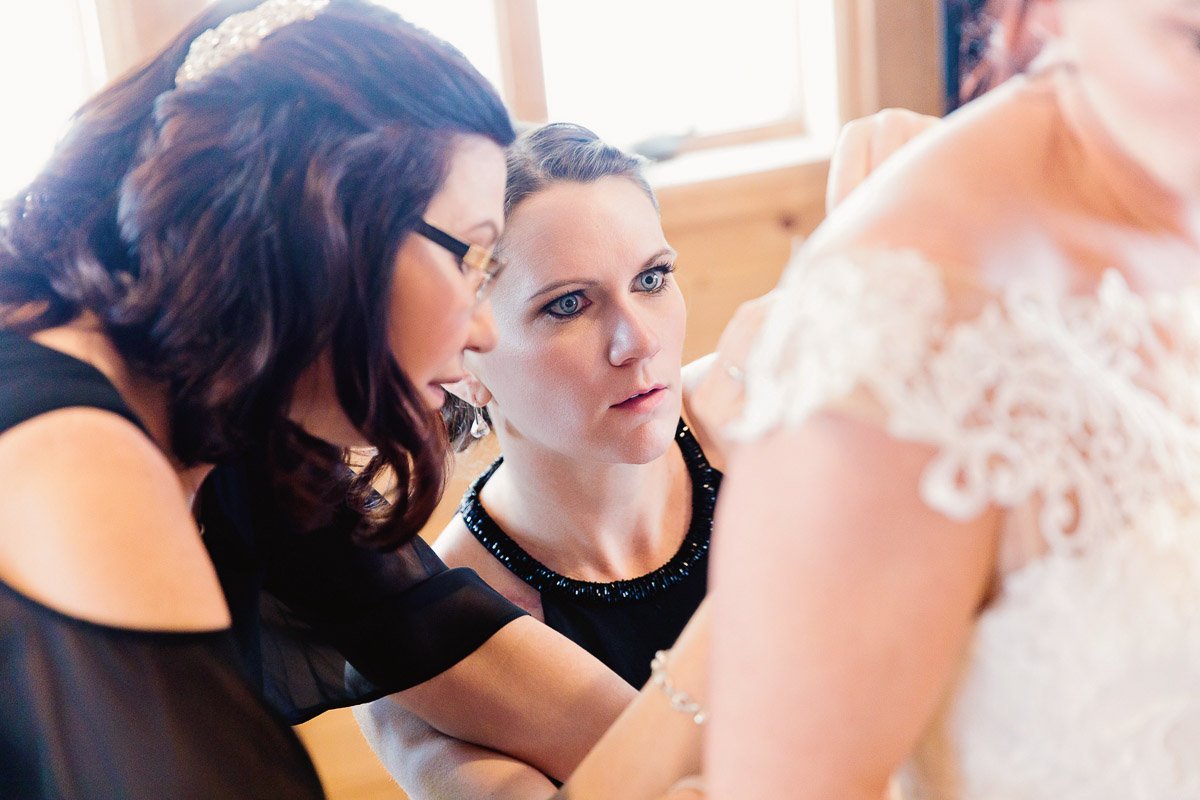Two women focus intently on adjusting a bride's dress in a well-lit room. The atmosphere is concentrated and supportive, highlighting teamwork.