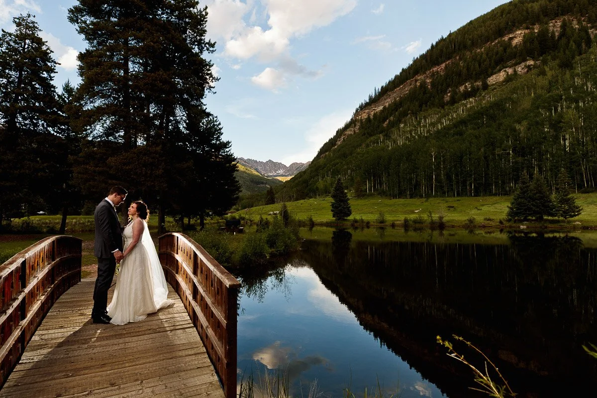 A bride and groom stand closely on a wooden bridge over a serene lake, surrounded by lush trees and mountains under a clear blue sky. Romantic and tranquil.