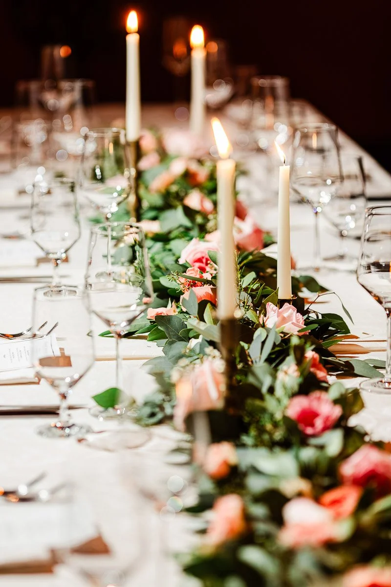 Elegant table setting with lit candles and a floral centerpiece of pink roses and greenery. Crystal glasses and fine dining ware create an intimate, refined ambiance.