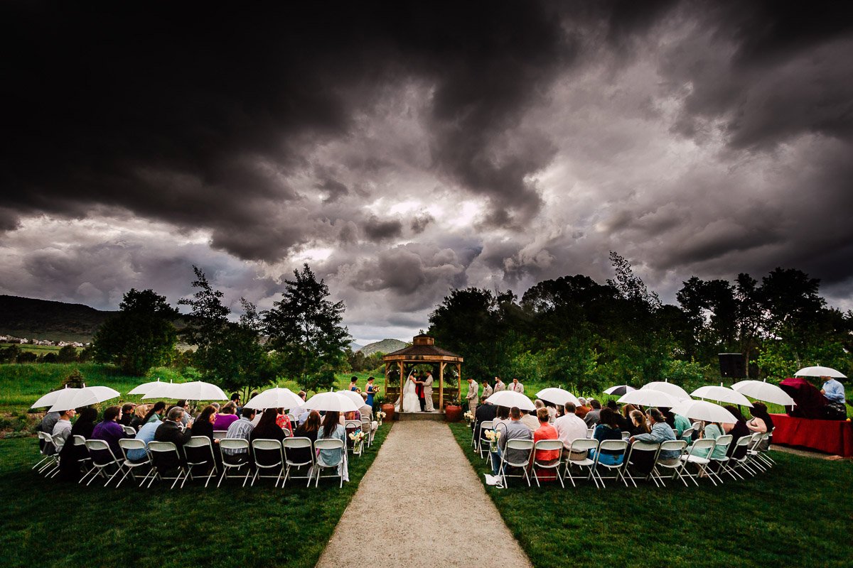 Outdoor wedding with dark storm clouds overhead. Guests seated in colorful chairs hold white umbrellas, focusing on a couple at a wooden altar. Dramatic mood during a Chatfield Farms wedding in Littleton, Colorado.