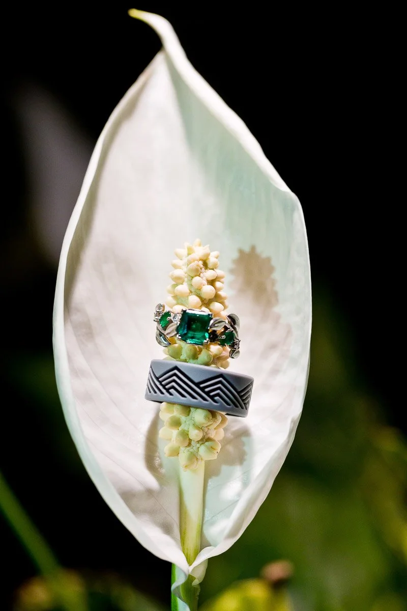 Two rings rest on a white peace lily stamen. The top ring features a vibrant green gemstone, while the bottom ring has a black zigzag pattern.