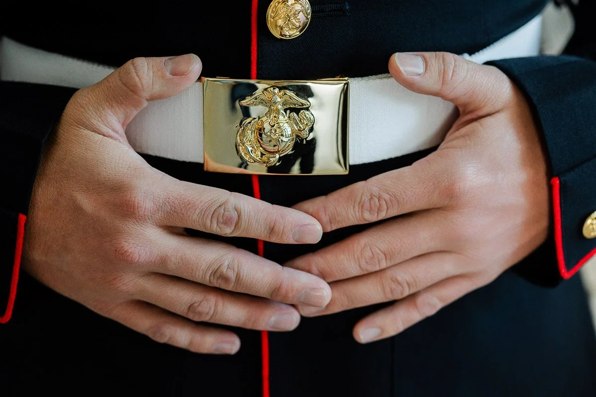Close-up of a person wearing a formal Marine uniform with gold buttons. Hands rest over a shiny gold eagle emblem on a white belt, conveying pride.