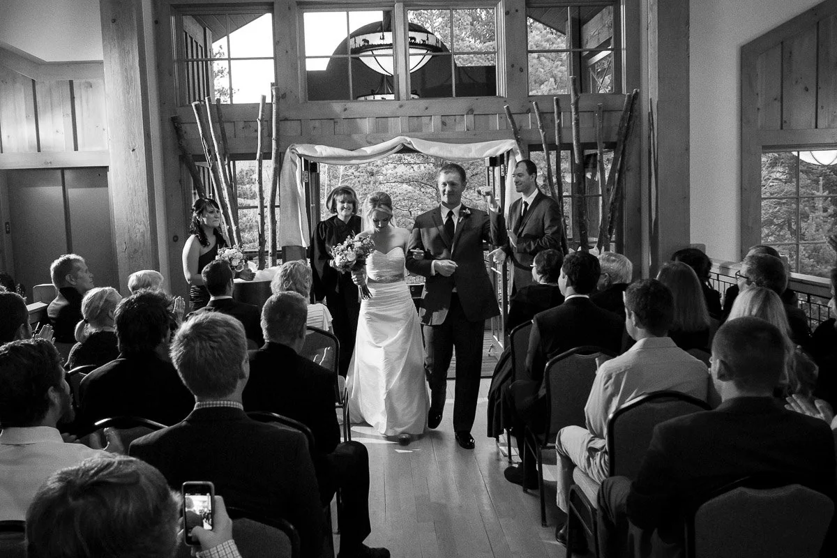A bride in a white gown and a groom in a suit walk down the aisle together, smiling. Guests sit on either side, and large windows fill the room with light.