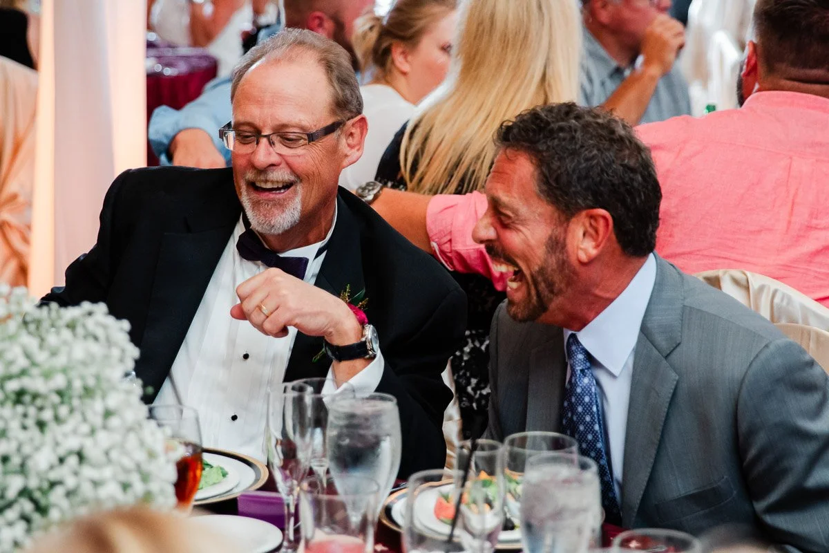 Two men in suits share a joyful laugh at a formal event, surrounded by dining tables and guests. The mood is lively and festive.