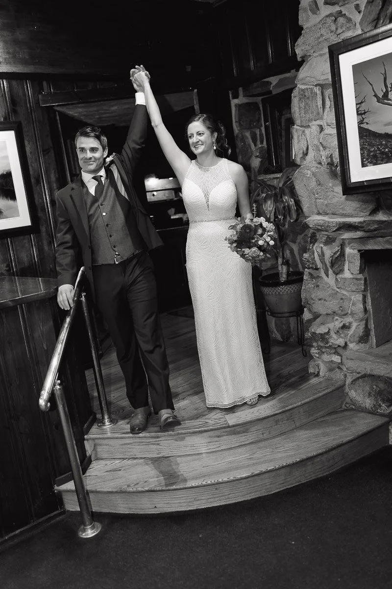 Bride and groom joyfully enter a rustic venue, holding hands high. Bride wears a lace gown, groom in a suit. Mood is celebratory and elegant.