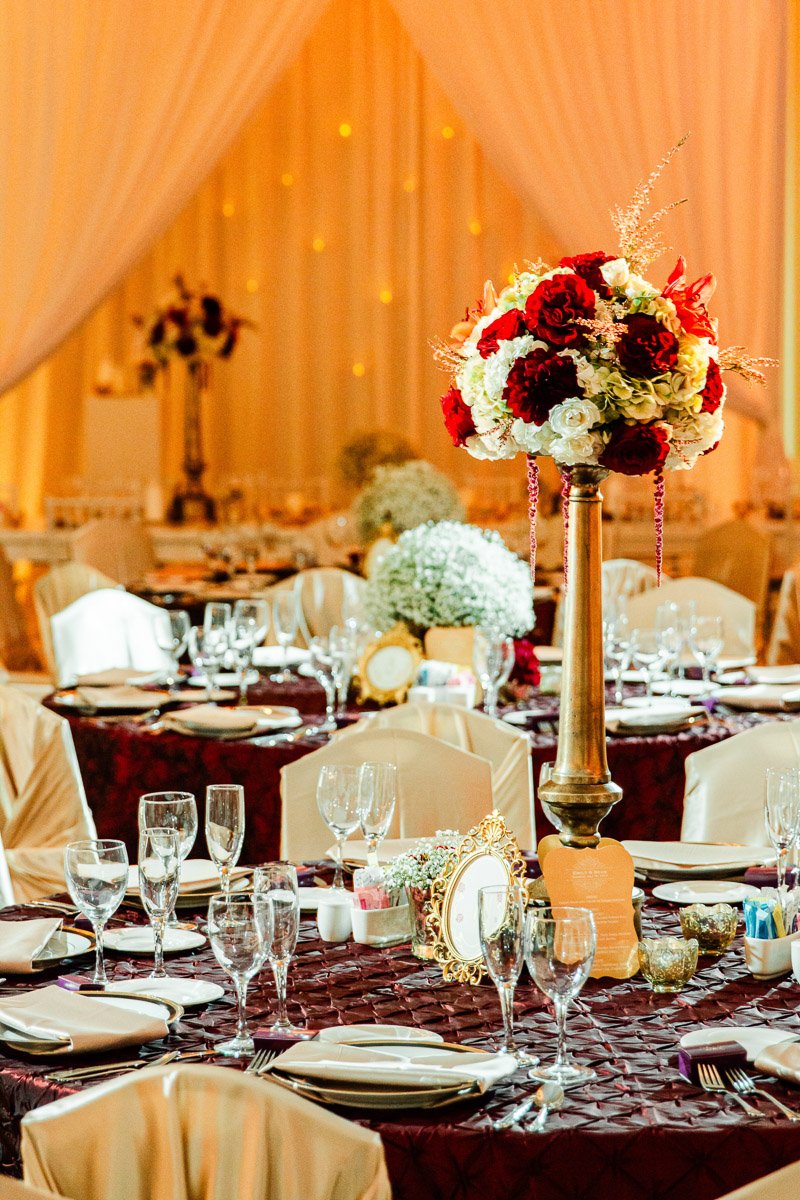Elegant wedding reception with tables draped in maroon cloths, adorned with tall floral centerpieces featuring red and white flowers. Warm lighting creates a romantic ambiance.