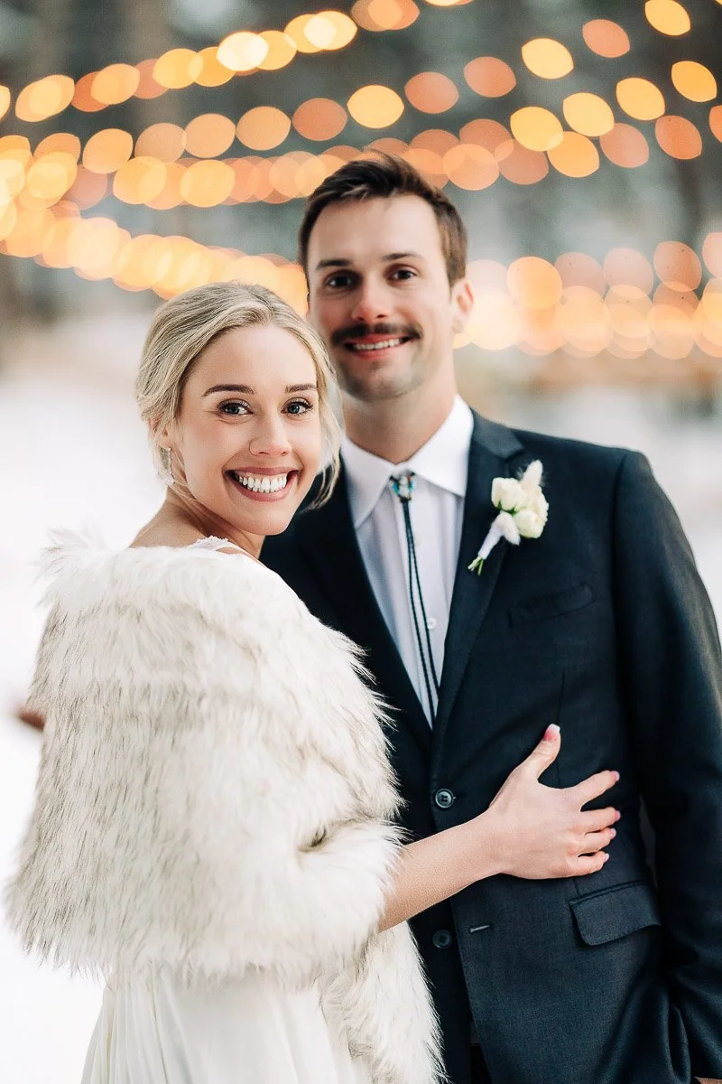 A joyous bride and groom stand together outdoors; the bride in a fur stole, the groom in a suit with a floral boutonniere, beneath warm, glowing string lights.