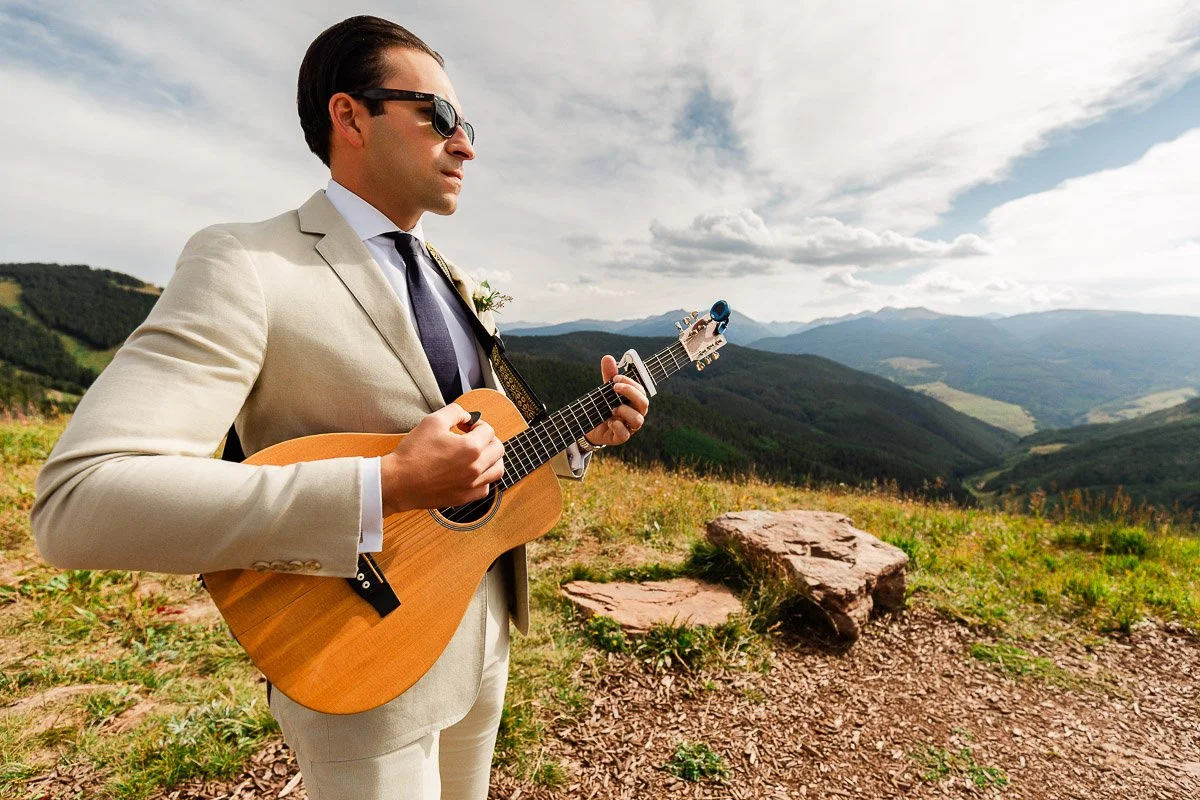 A man in a light suit and sunglasses plays an acoustic guitar in a picturesque mountain setting. The sky is partly cloudy, evoking a serene and contemplative mood.