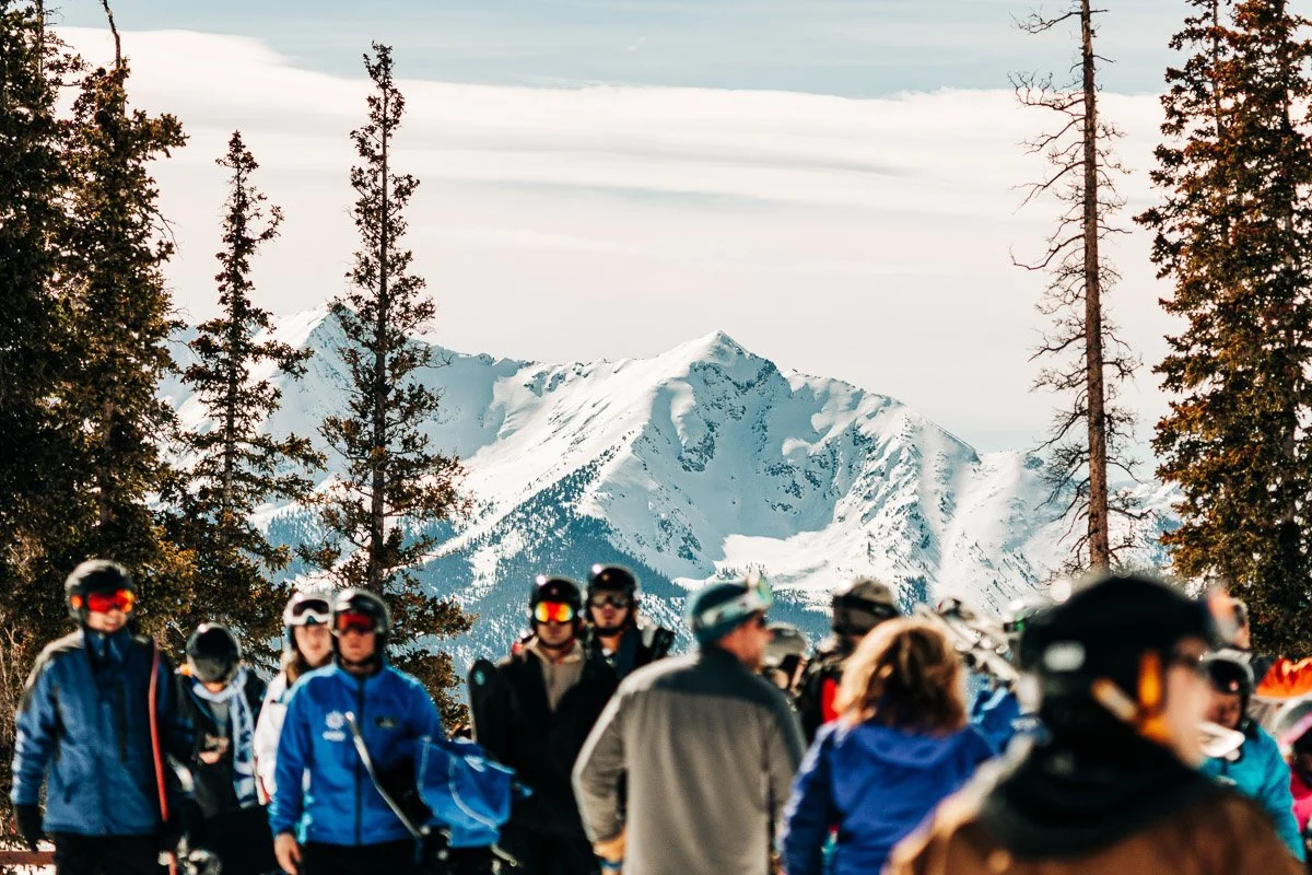 A group of skiers in colorful gear and helmets gather on a snowy mountain with a backdrop of snow-covered peaks. Tall pine trees frame the scene.