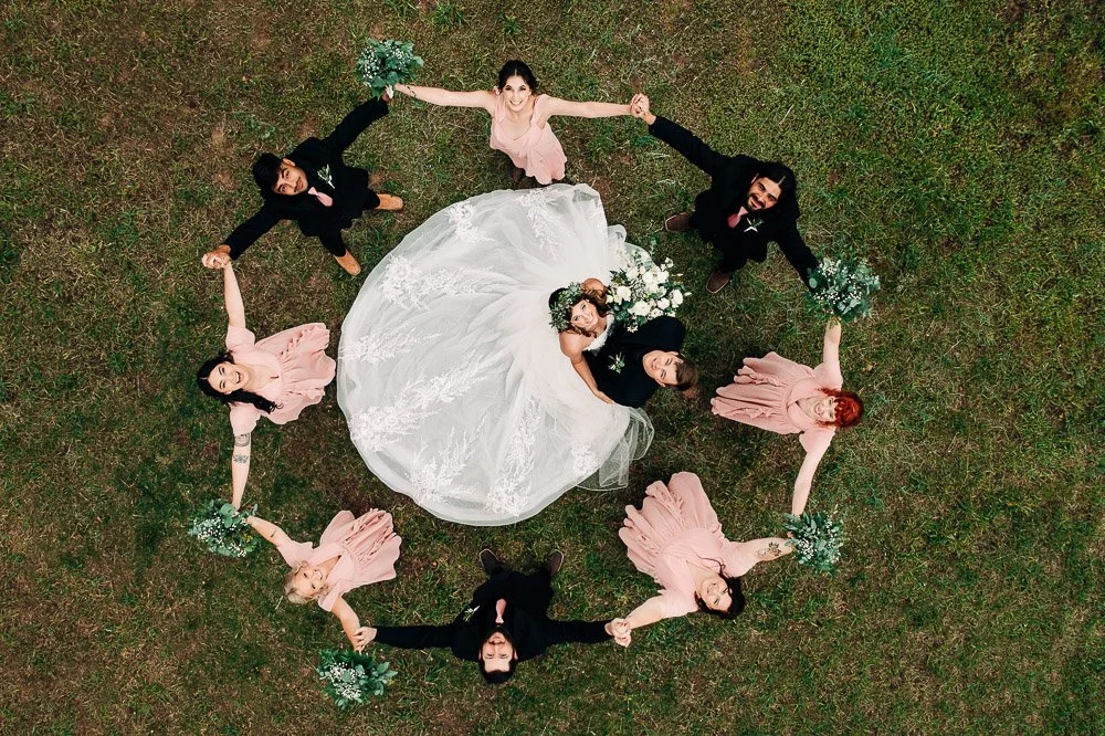 Aerial view of a wedding party forming a circle on grass. The bride and groom lie in the center, surrounded by happy bridesmaids and groomsmen holding hands.