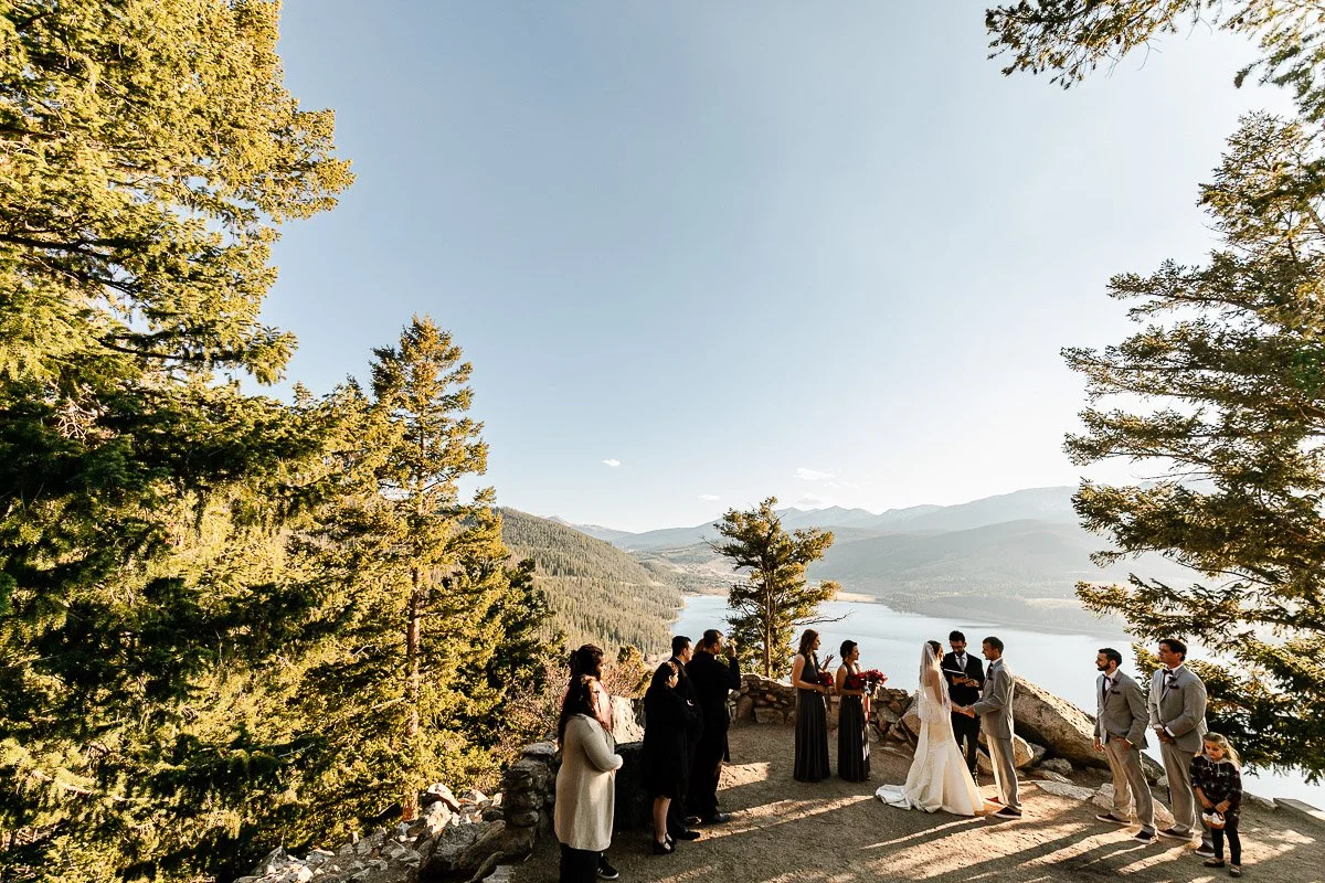 A wedding ceremony takes place outdoors on a cliffside overlooking a lake and mountainous landscape. Guests stand around the couple amid tall pine trees.