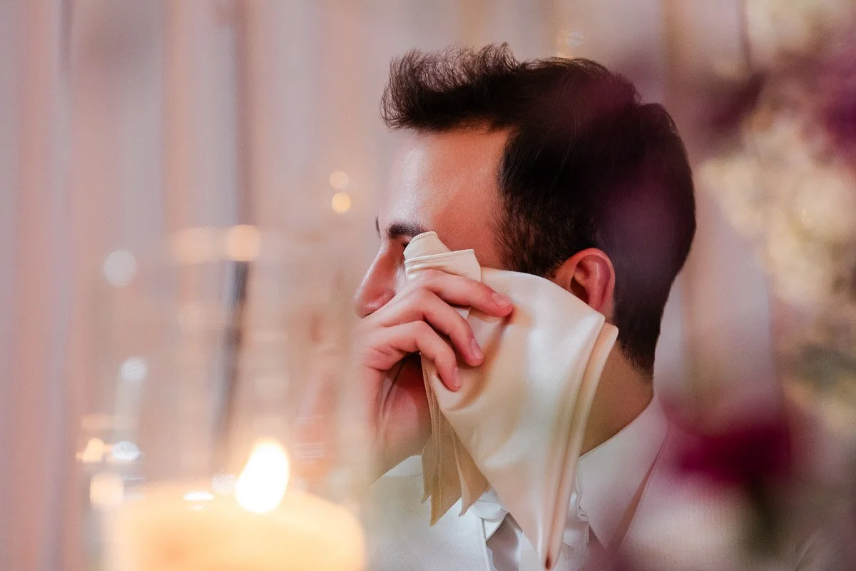 A man in a suit wipes tears with a handkerchief at an emotional event. The warm glow of a candle and soft focus adds a heartfelt tone.