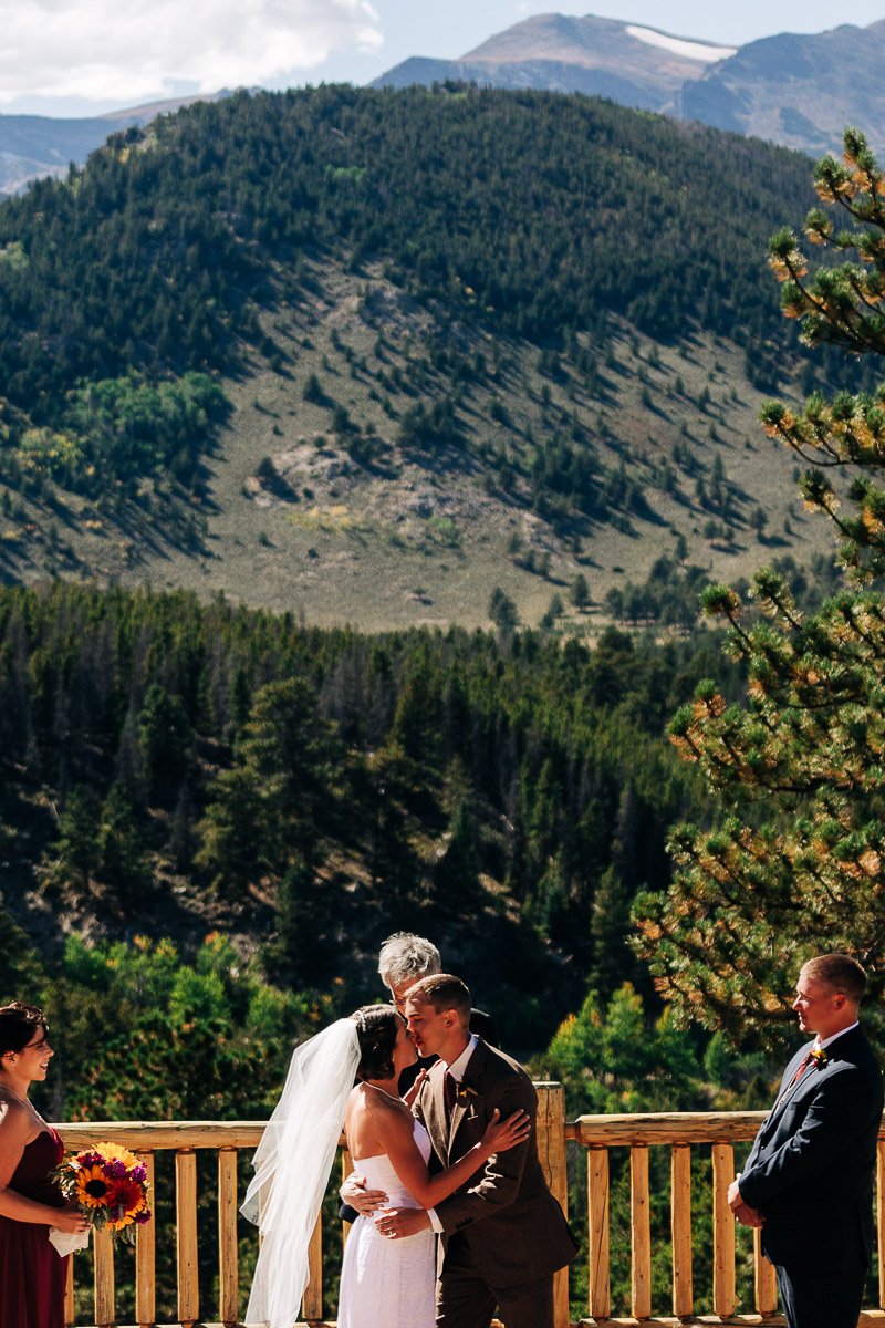 A couple shares a kiss at their outdoor wedding, set against a backdrop of lush green mountains. Bridesmaid holds flowers on the left, officiant and groomsman on the right.
