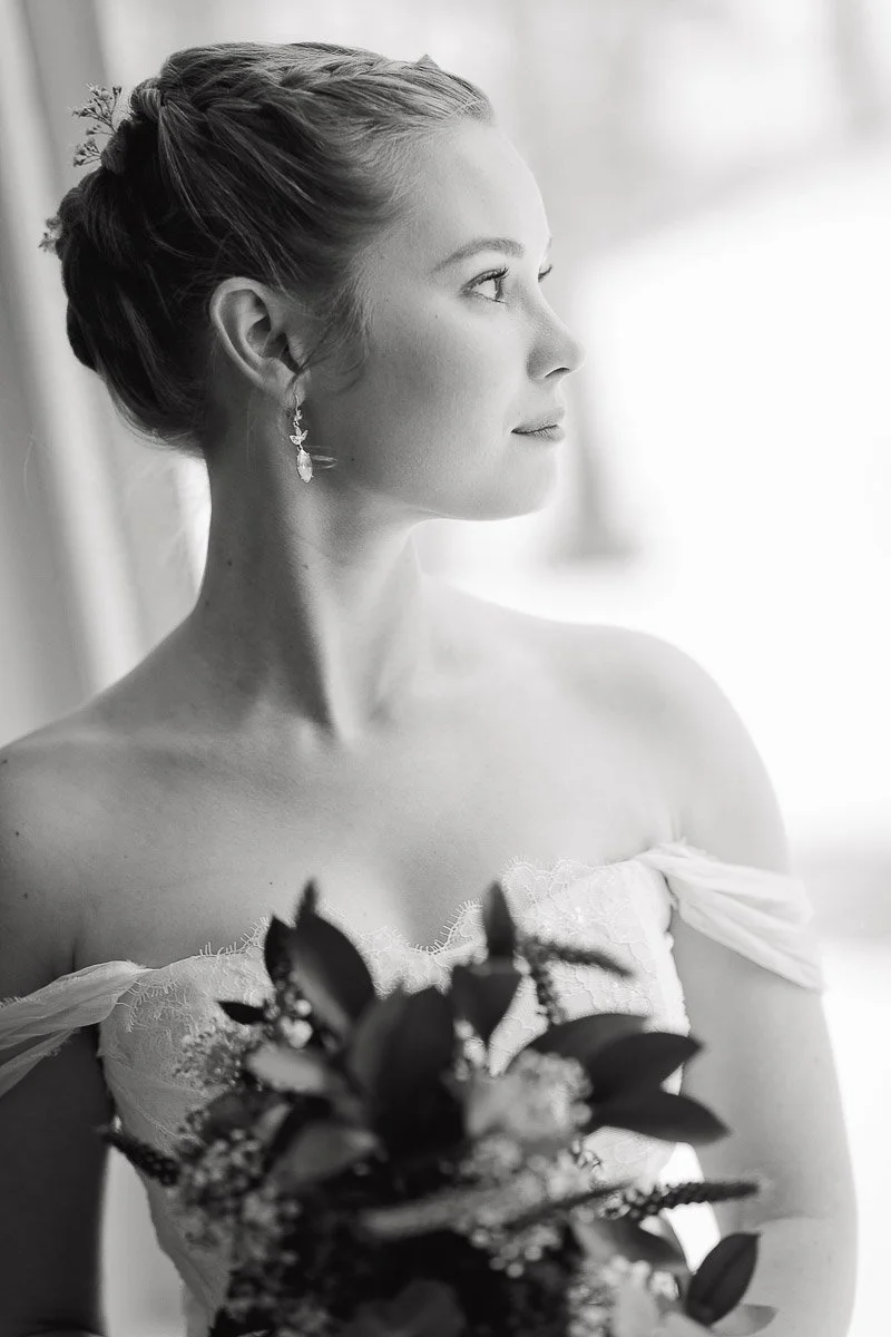 Black and white photo of a bride in an off-shoulder dress holding a bouquet, gazing thoughtfully to the side, with soft lighting and a serene expression.