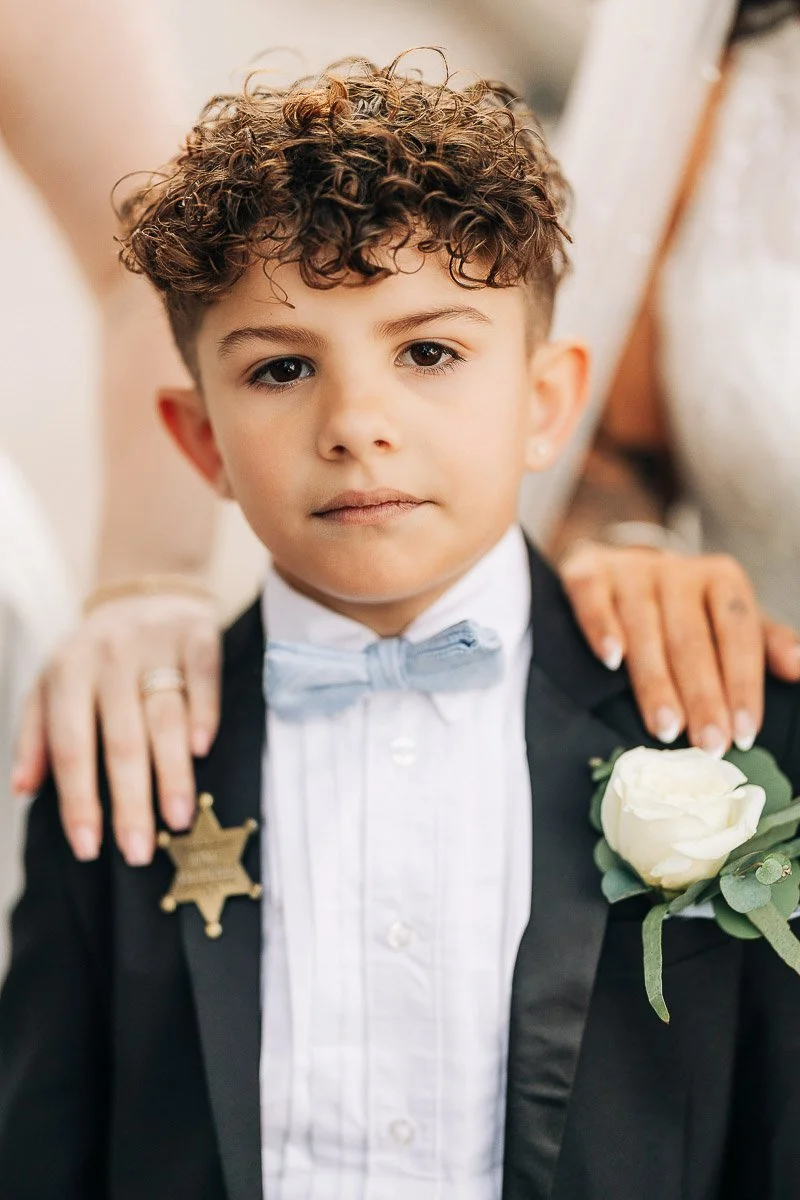 Young boy in a suit with a light blue bow tie and white shirt, wearing a badge and a white rose boutonniere. Hands gently rest on his shoulders.