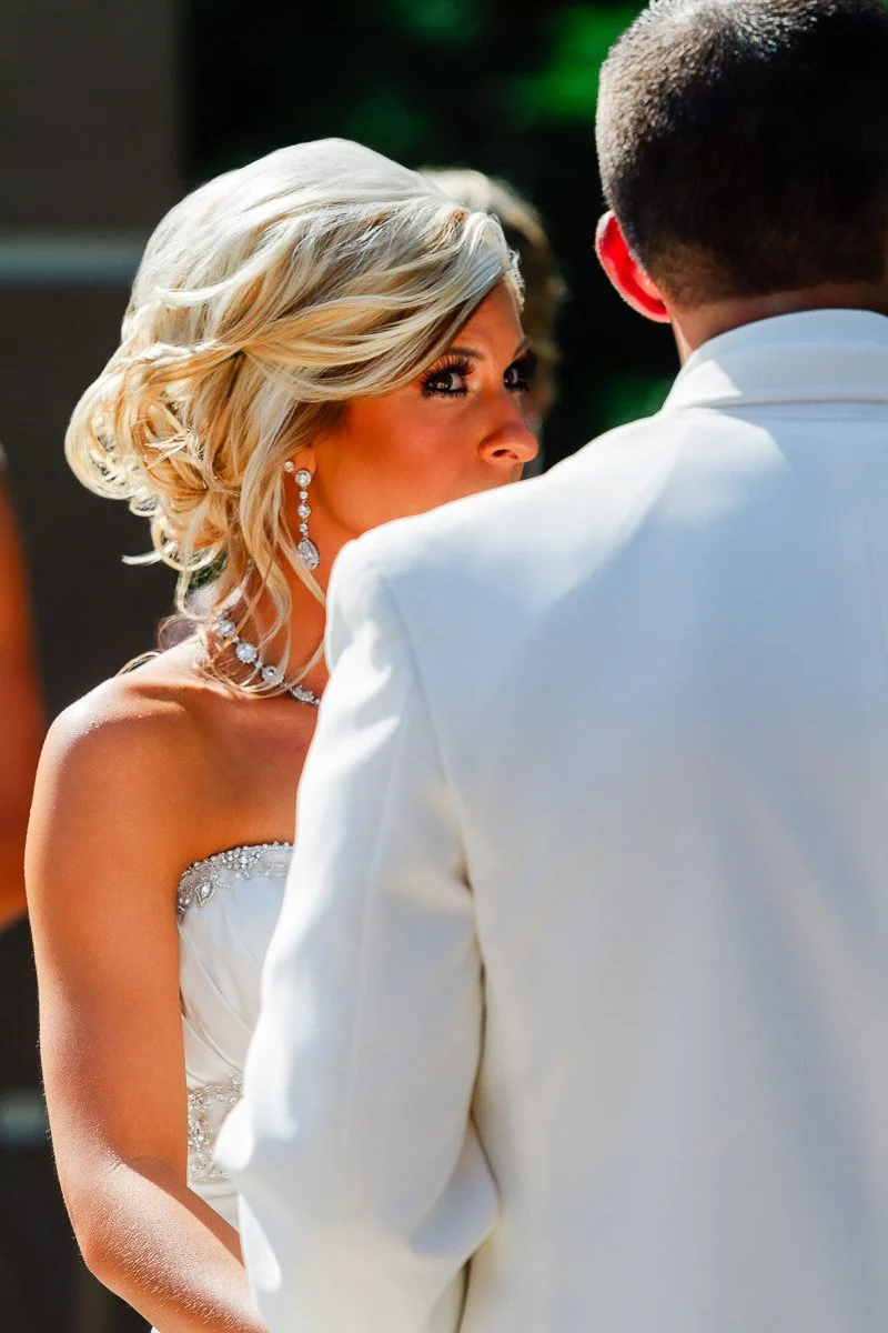 A bride with blonde hair and jeweled earrings looks intently at the groom during an outdoor ceremony. Both wear elegant white attire, conveying a romantic tone.