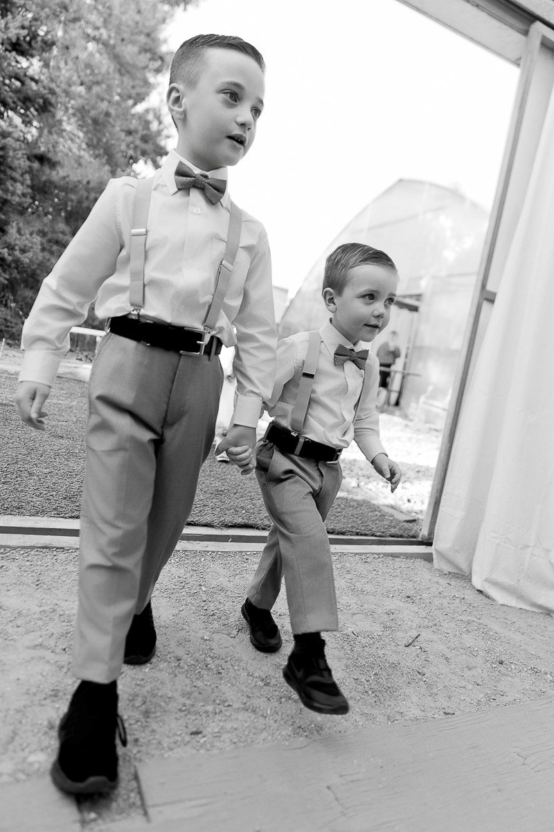 Two young boys in matching outfits, with suspenders and bow ties, walk hand-in-hand down an aisle. They look determined and joyful.
