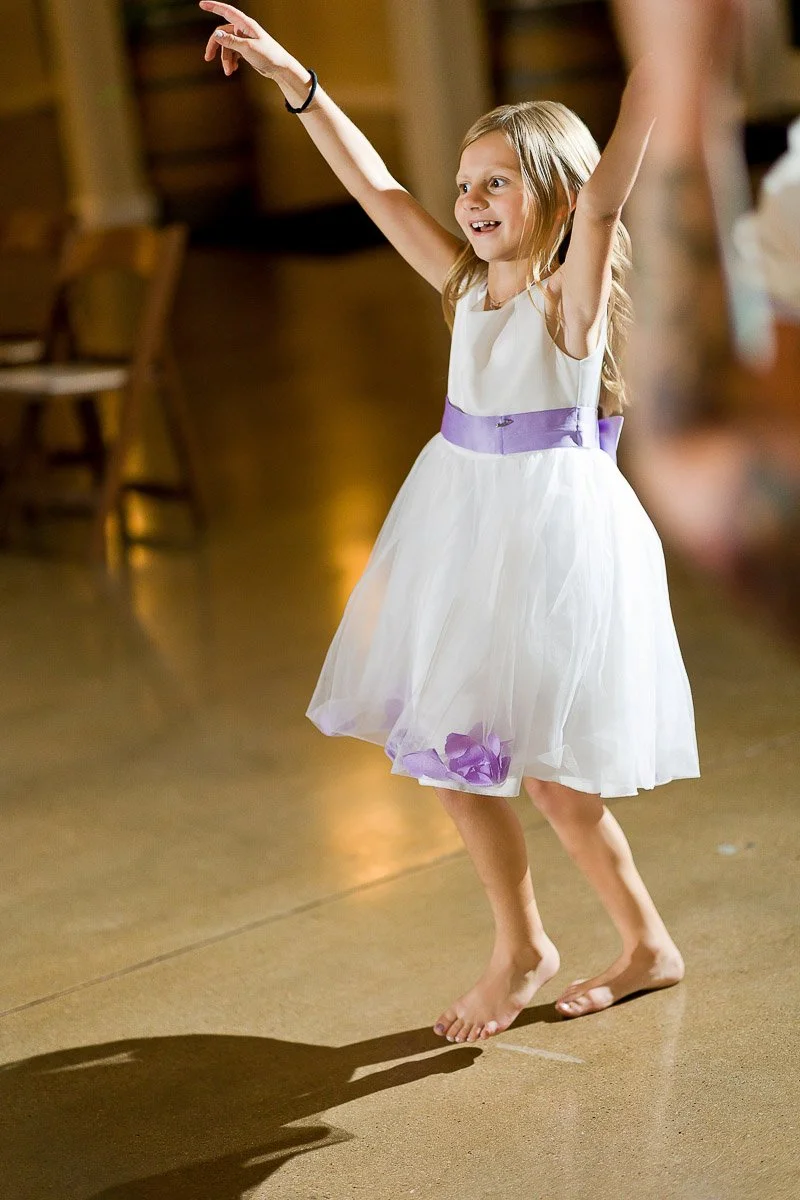 A joyful young girl in a white dress with a purple ribbon dances barefoot on a polished floor. Her arms are raised, and she smiles brightly.