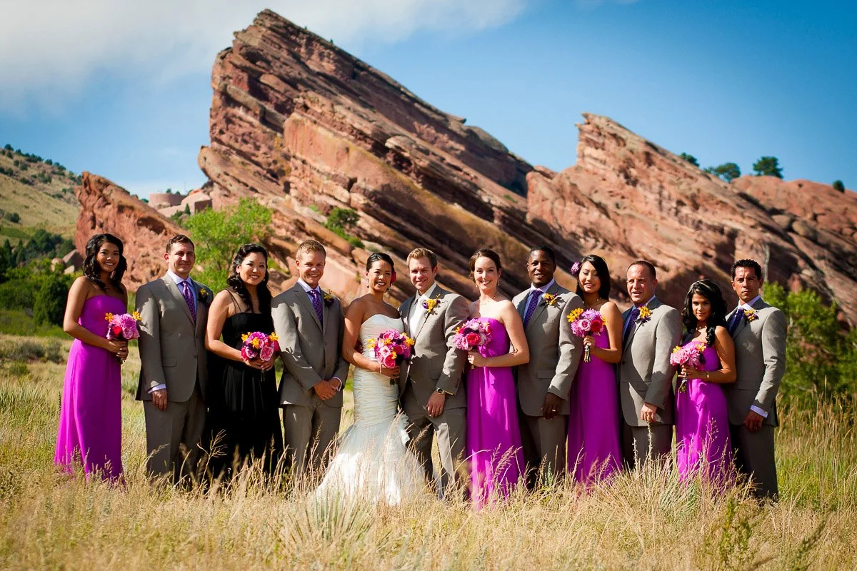 A wedding party poses joyfully outdoors. The bride and bridesmaids in purple dresses hold bright bouquets. Groomsmen in grey suits stand against stunning red rock formations captured by Red Rocks wedding photographer tomKphoto