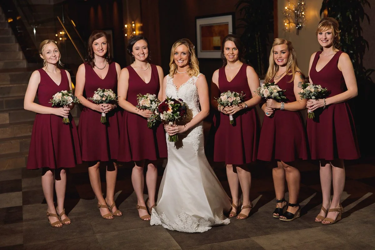 Bride in a white gown stands with six bridesmaids in burgundy dresses. Each holds a bouquet, smiling in an elegant, warmly lit indoor setting.