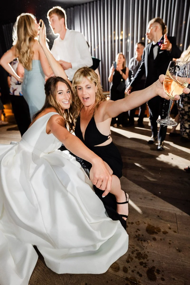 A joyful bride in a white dress and a woman in black are seated on the dance floor, raising their arms and smiling. Around them, people are dancing.