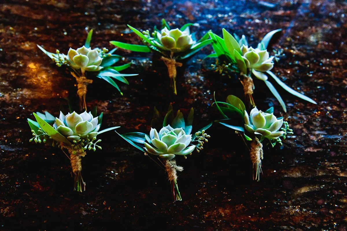 Six small succulent boutonnieres with greenery, tied with twine, are arranged on a textured, dark marble surface. The mood is earthy and elegant.