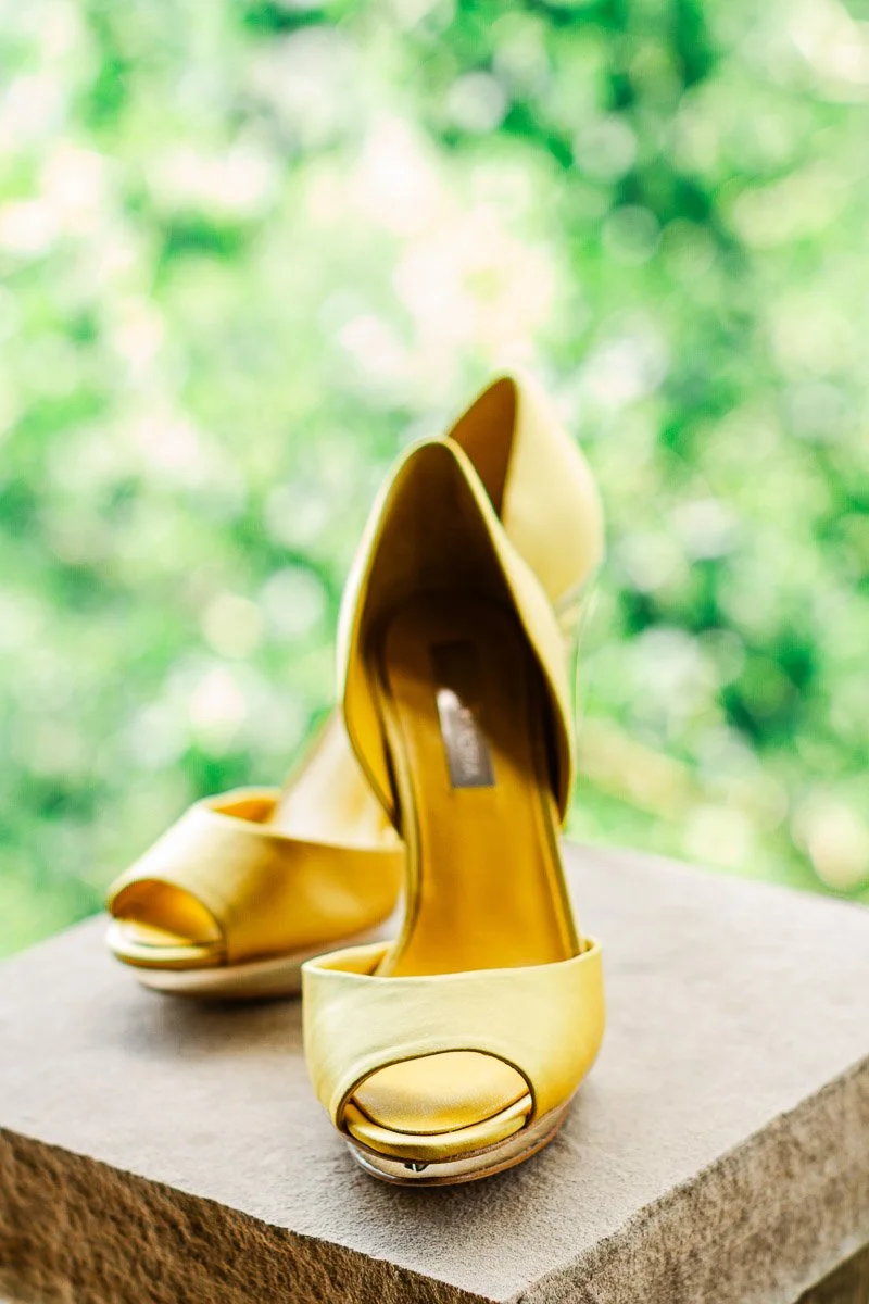 A pair of elegant, golden peep-toe high heels sit on a stone surface against a blurred, vibrant green background, evoking a sense of luxury and style.