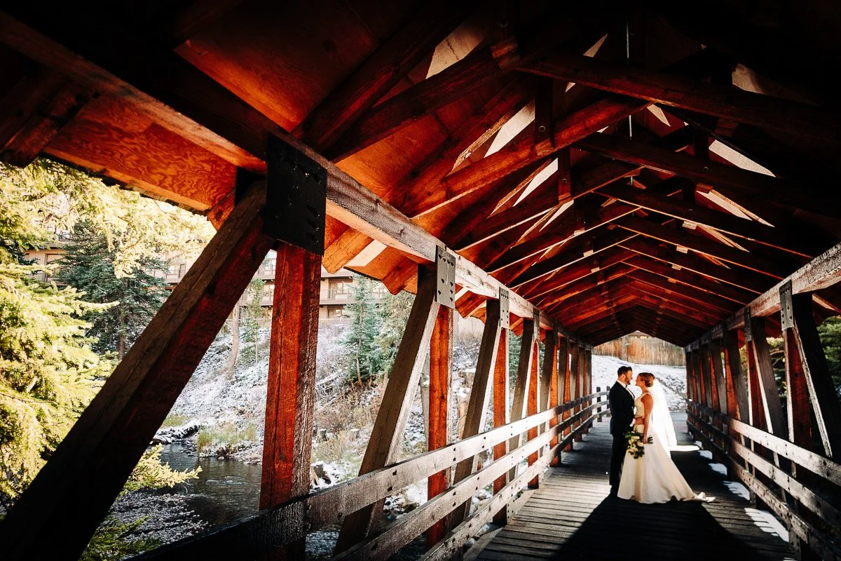 A bride and groom stand closely on a wooden covered bridge with sunlit beams. Snow-dusted trees and a peaceful river are visible in the background.