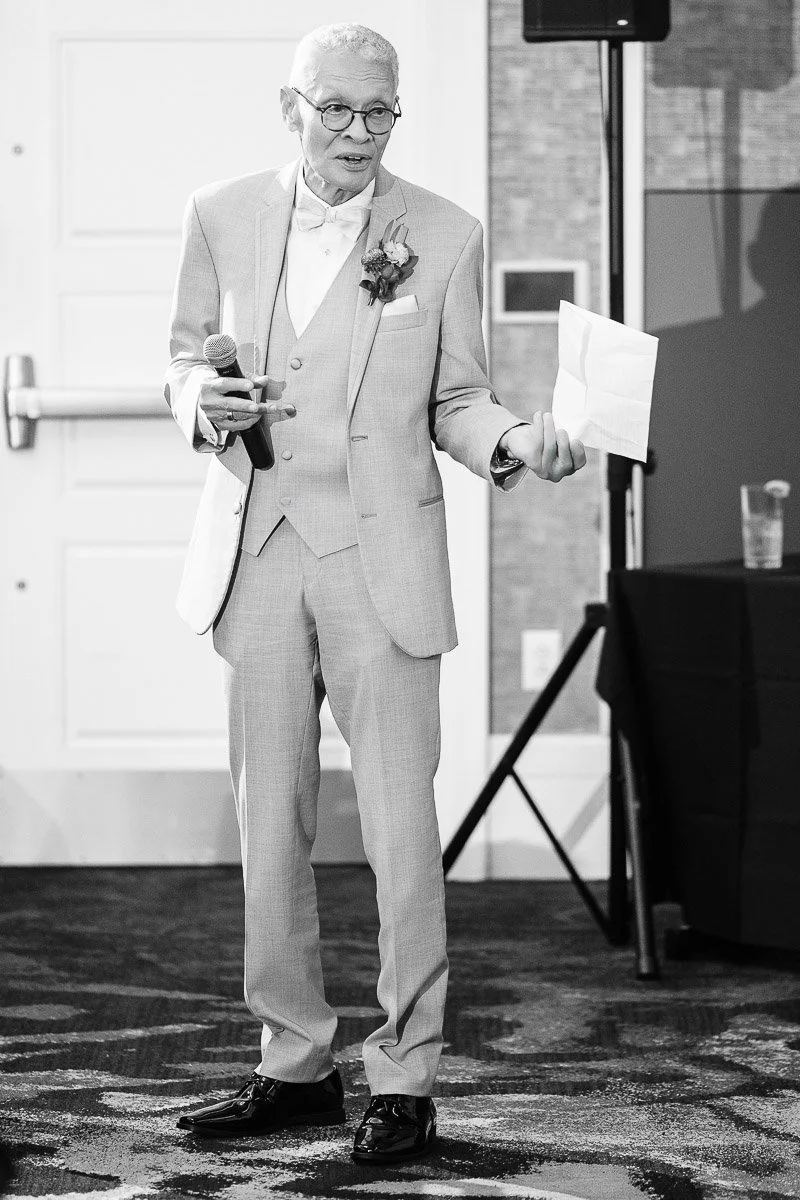 Elderly man in glasses and light suit gives a speech at an event, holding a microphone and paper. The setting is formal and he appears thoughtful.
