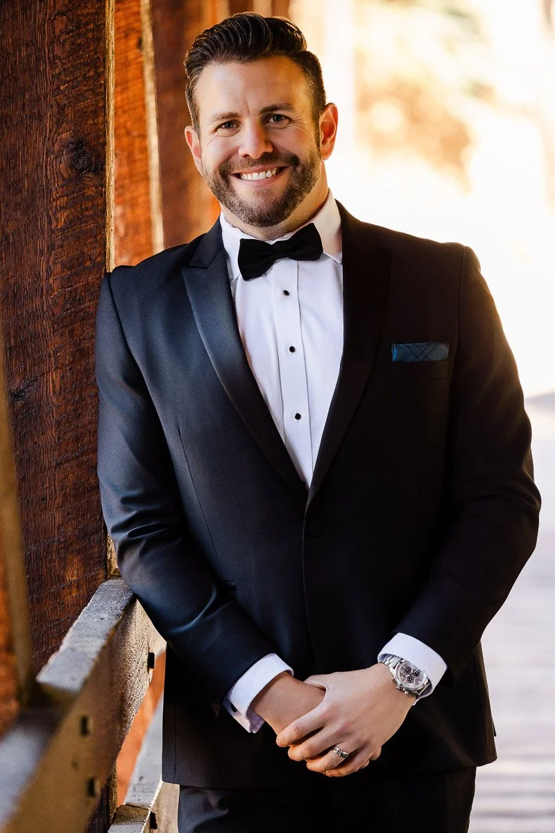 Man in a black tuxedo with a bow tie smiles warmly while leaning against a wooden railing. Sunlight casts a soft glow, creating a formal, cheerful tone.