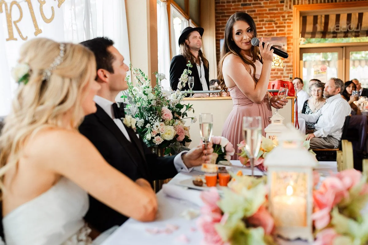 A woman in a pink dress gives a speech at a wedding reception. A seated bride and groom smile, holding champagne. Floral decor and warm lighting enhance the joyful atmosphere.