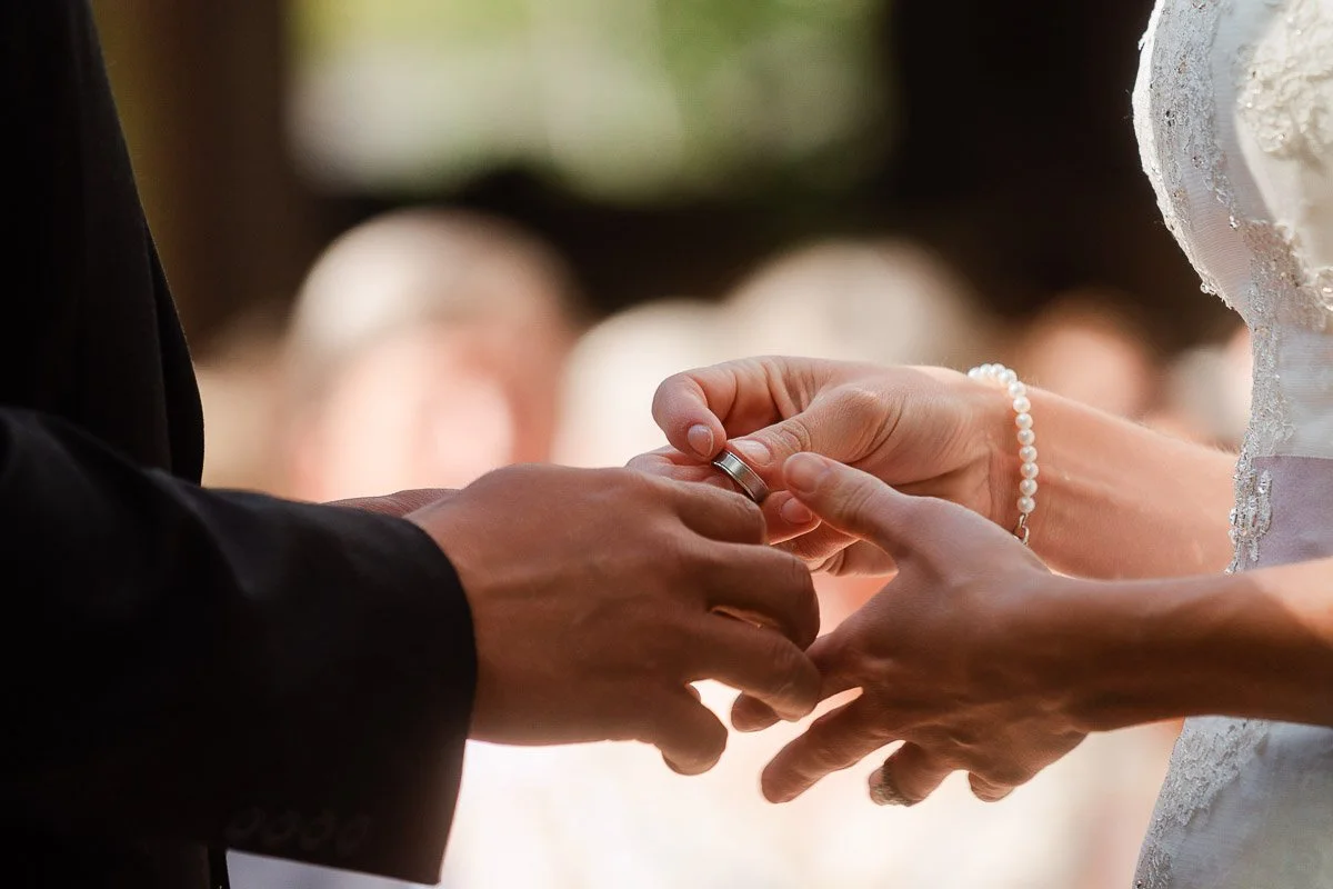 Close-up of a bride placing a wedding ring on the groom's finger during a ceremony. The scene conveys love and commitment, with soft focus in the background.