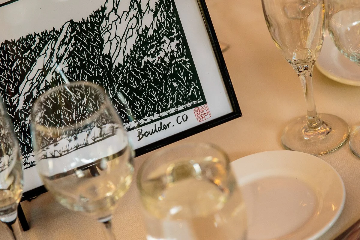 Framed illustration of Boulder, CO mountains on a dining table with wine glasses. The setting feels elegant and sophisticated.