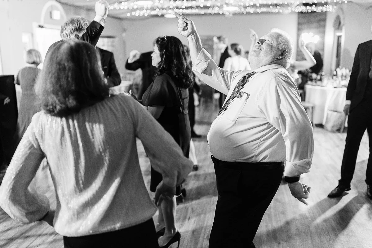 Black and white image of joyful people dancing at an event, with one man energetically pointing upward. The mood is festive and celebratory.