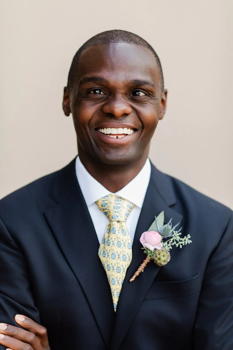 Smiling groom in a navy suit with a yellow patterned tie and floral boutonniere, exuding joy against a soft beige background.