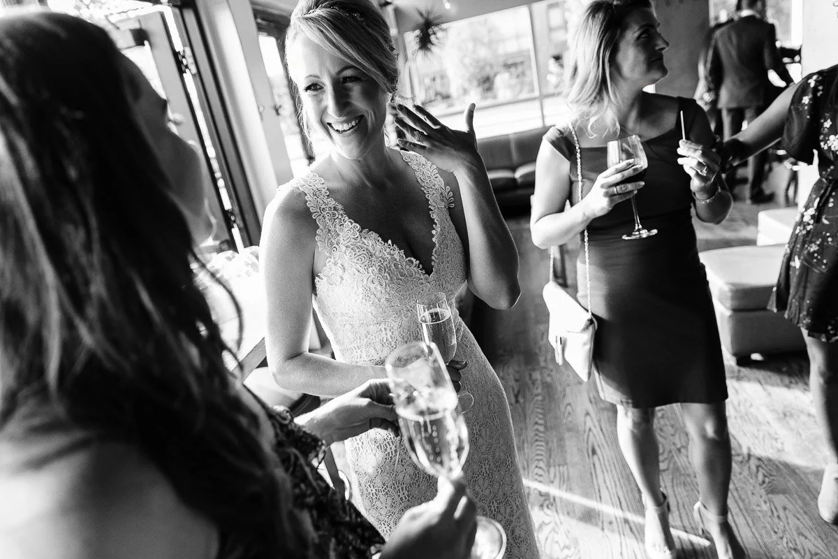A joyful bride in a lace dress holds a champagne flute, conversing with guests. The black-and-white photo captures a lively and celebratory atmosphere.