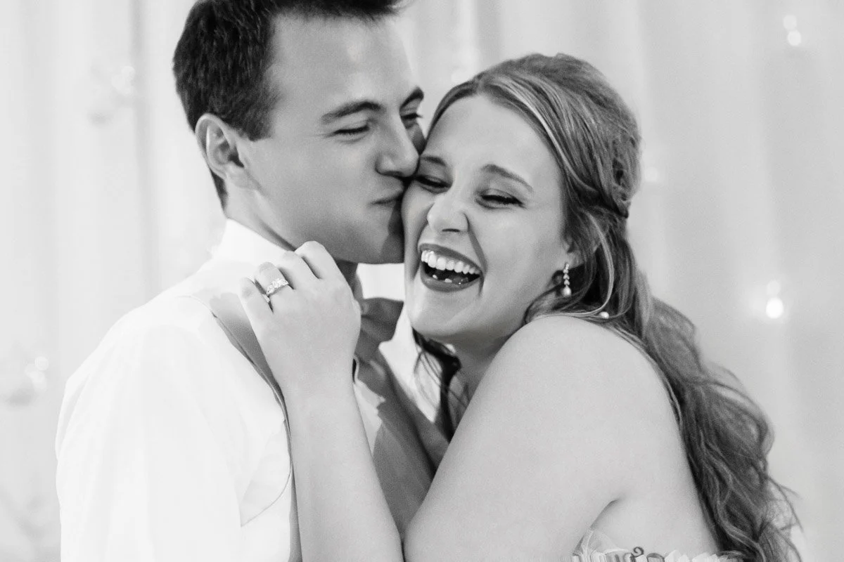A couple shares a joyful moment, with the man kissing the woman's cheek. She laughs with her eyes closed. Their expressions convey love and happiness.