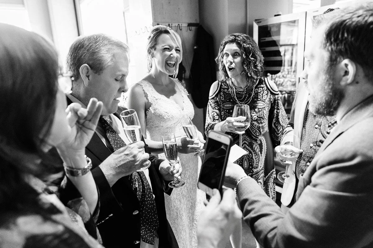 A joyous group gathers indoors, holding glasses and smiling. A woman in a wedding dress laughs, creating a cheerful and celebratory atmosphere. Black and white photo.