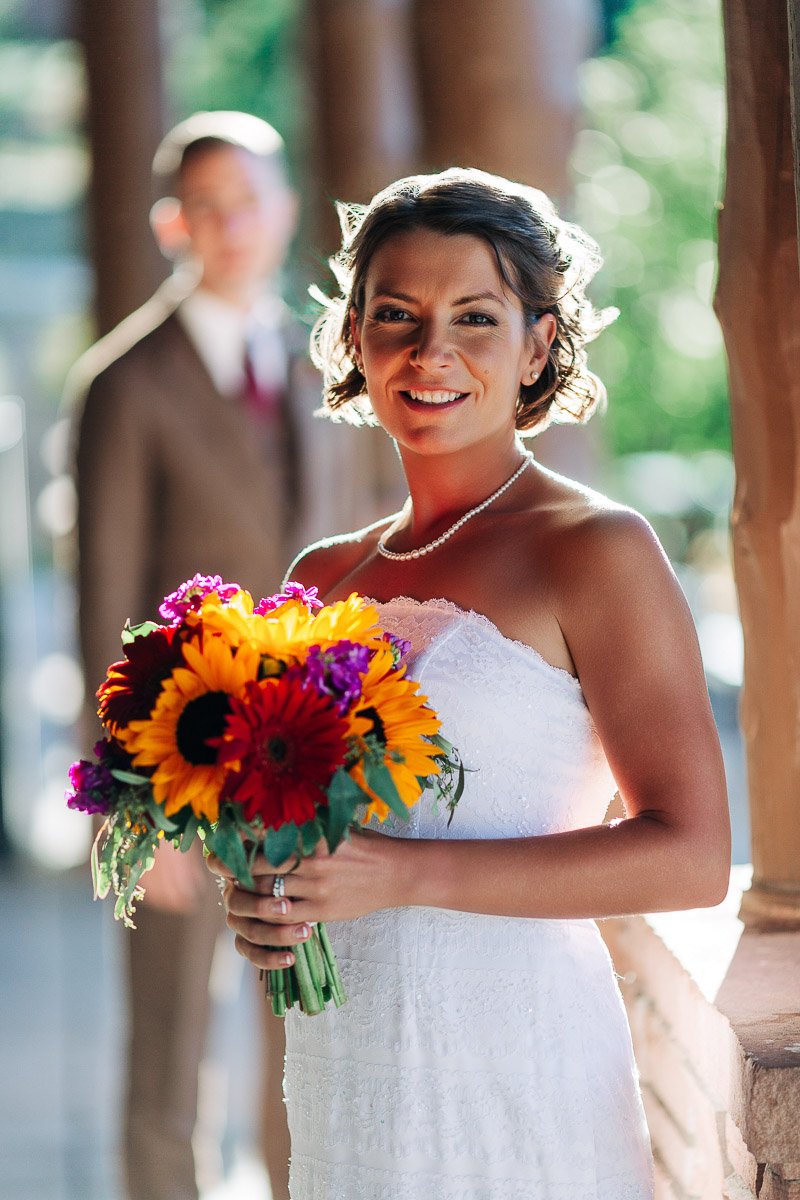 Bride in a white dress holding a vibrant bouquet of sunflowers and other colorful flowers, smiling warmly. A person in a suit stands blurred in the background.